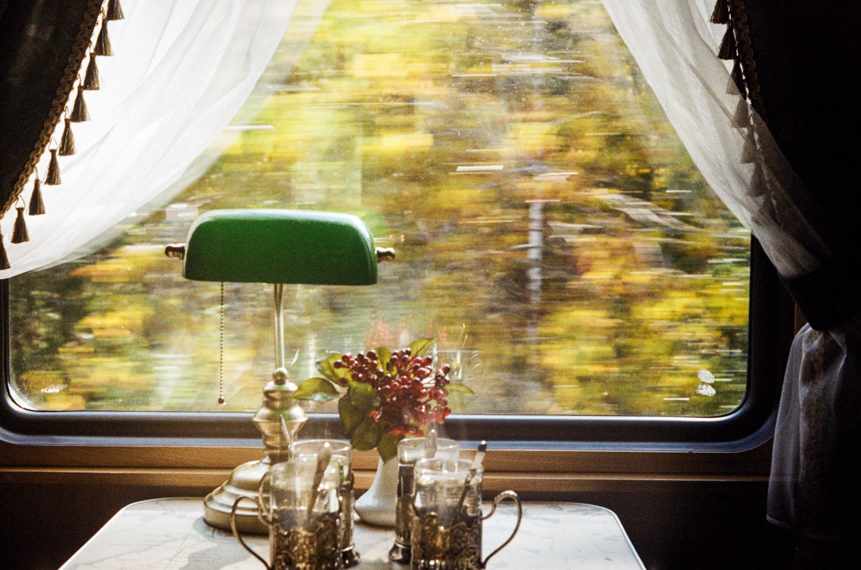 A brass lamp and white curtains in a train dining car, autumn trees blurring past the window